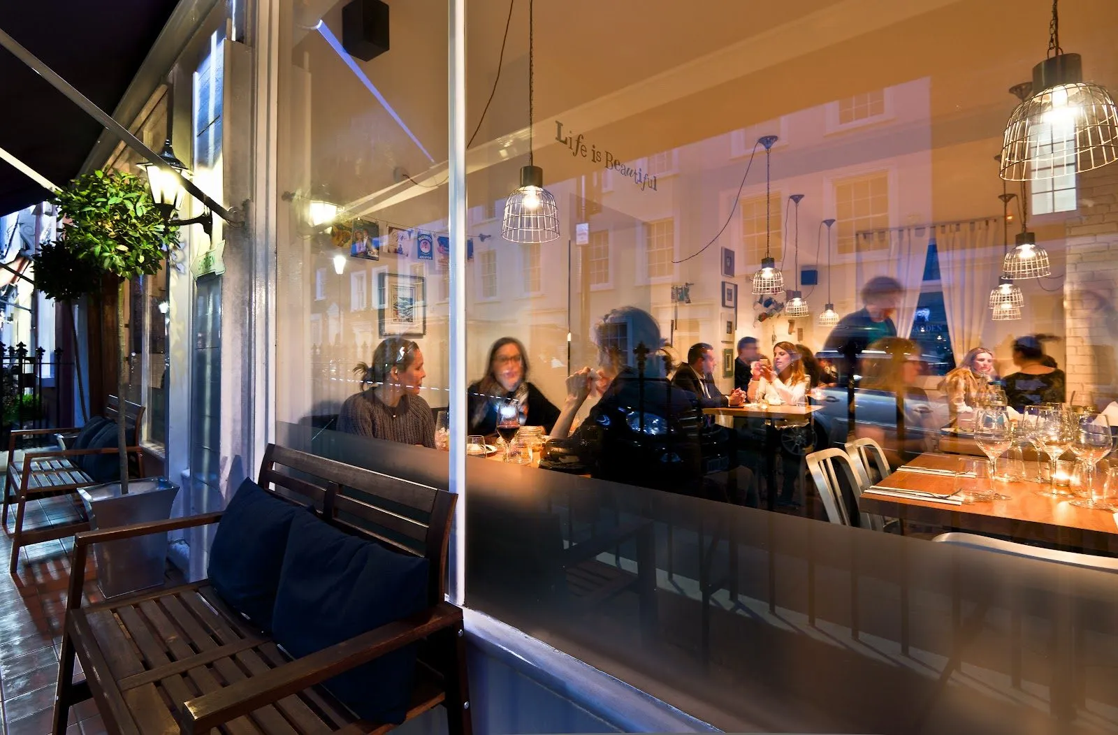 Diners at tables behind Mazi's front window in the evening, with wire cage pendant lights inside and a wooden bench on the terrace outside