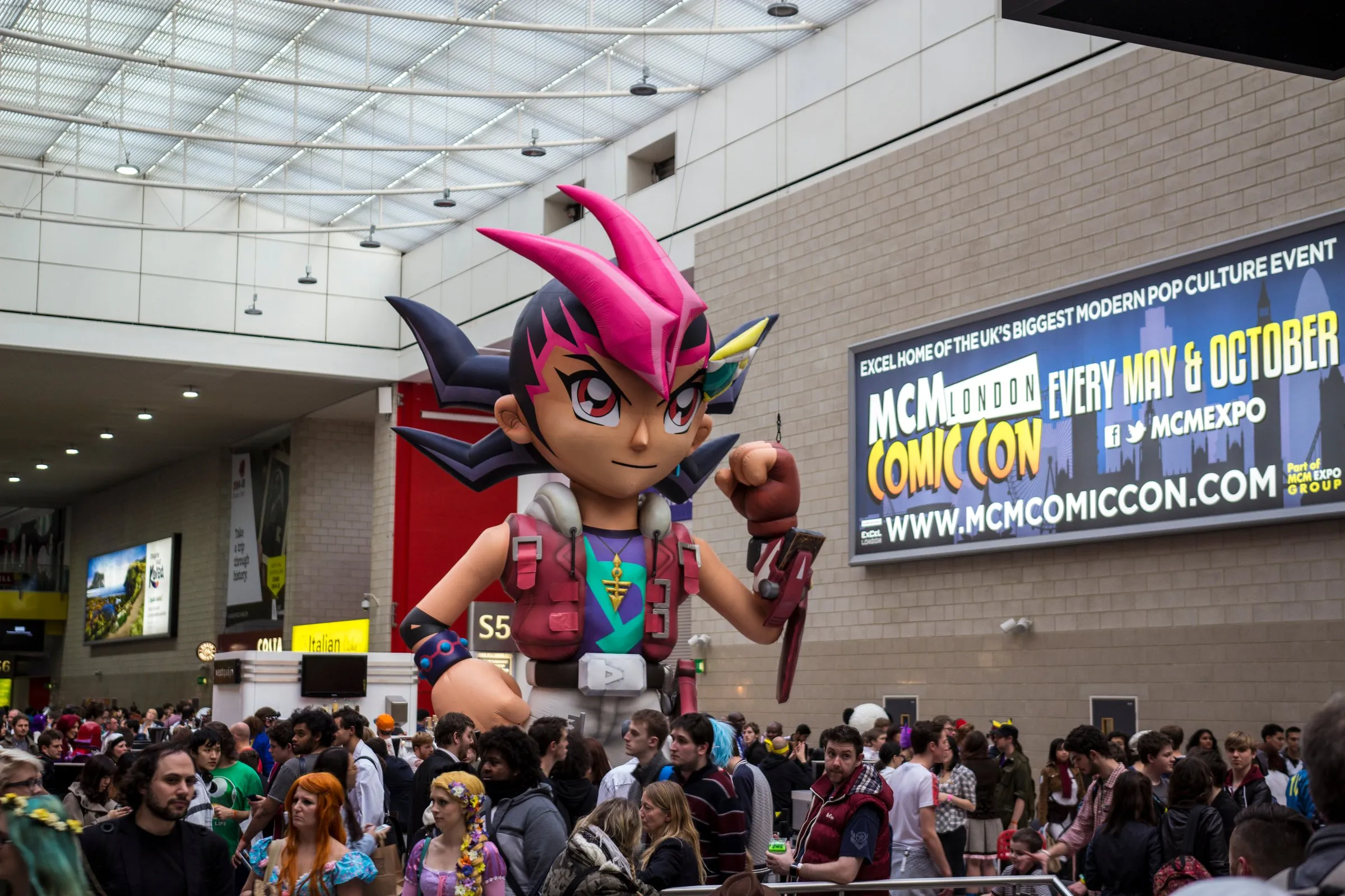A crowd on the show floor at MCM London Comic Con at ExCeL beneath a giant inflatable anime character, with the event's own wall sign reading 'MCM London Comic Con, every May & October'