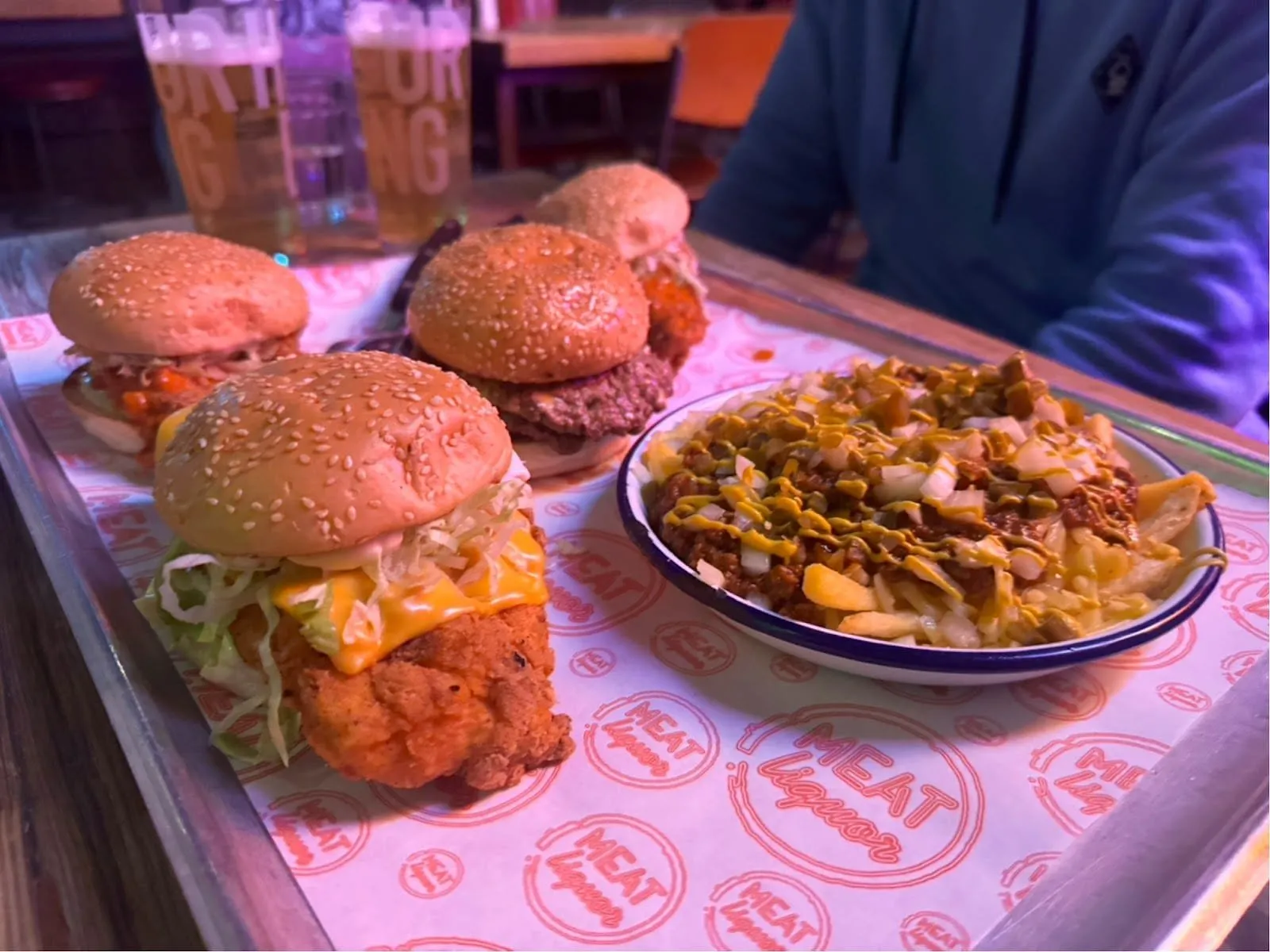 A MEATliquor tray lined with the restaurant's printed paper: a fried chicken burger with melted cheese and shredded lettuce, beef and chicken burgers in sesame buns behind it, and a bowl of chilli cheese fries with diced onion and mustard