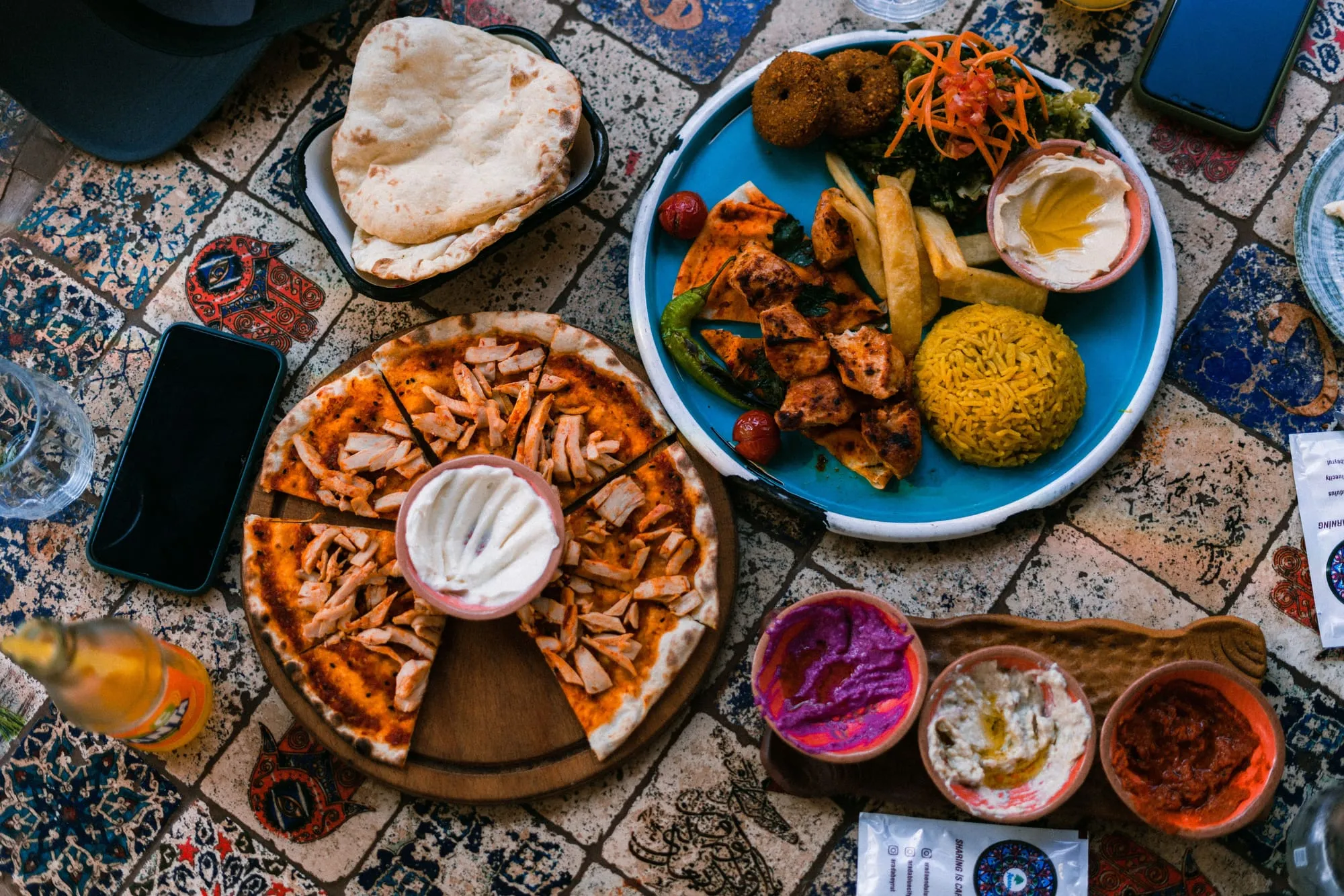 An overhead view of a shared mezze spread on a patterned tiled table - flatbreads, falafel, hummus, a sliced chicken flatbread and bowls of dips