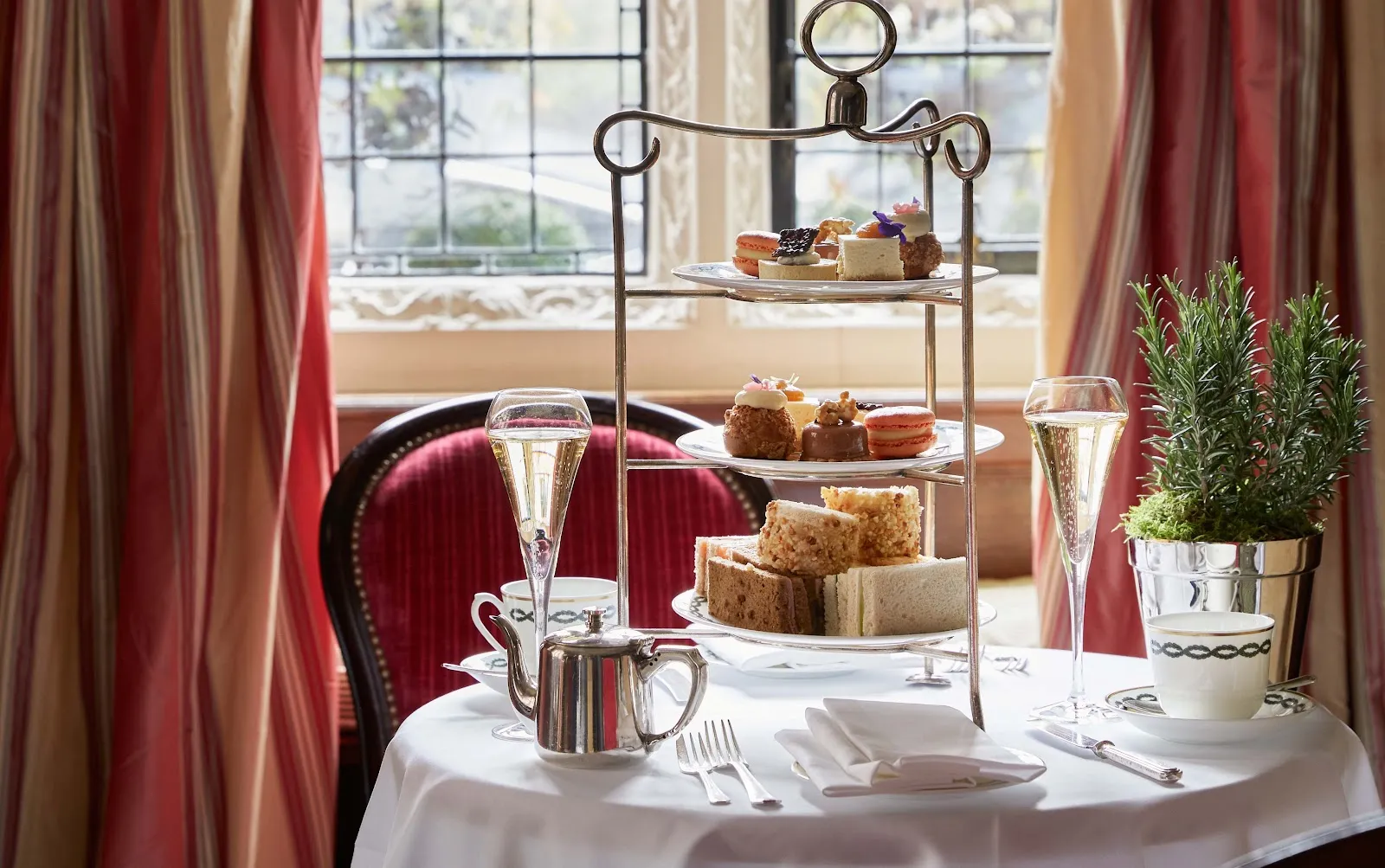 A three-tier afternoon tea stand on a white-clothed table at The Milestone, in front of a leaded window with striped curtains and a red velvet chair