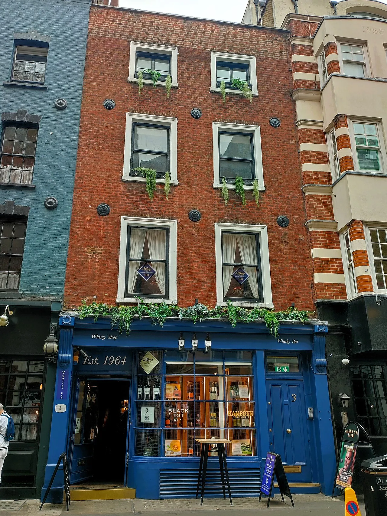 The blue shopfront of Milroy's of Soho on Greek Street, with Whisky Shop and Whisky Bar painted on the fascia and bottles in the window, under a four-storey brick house
