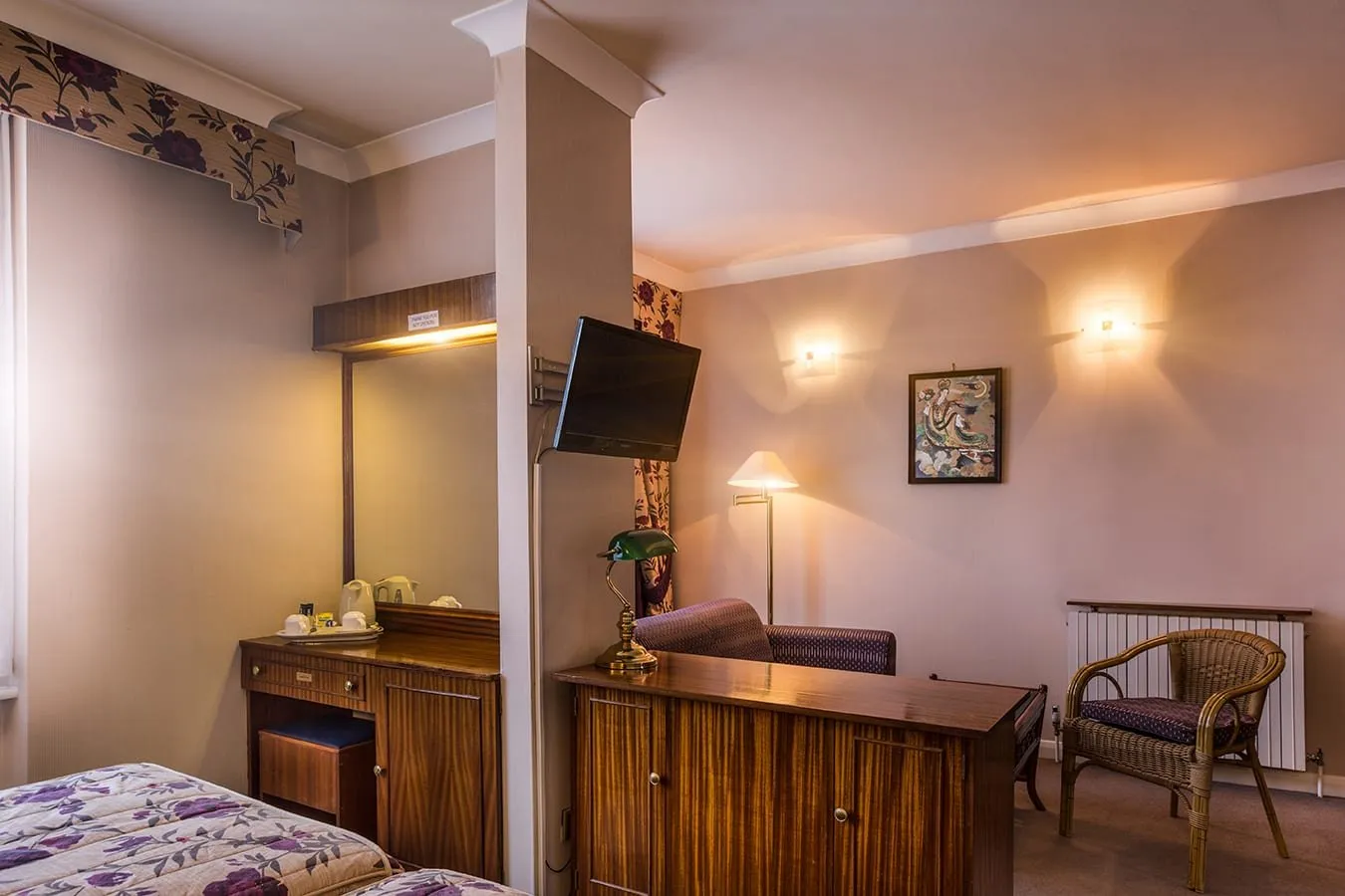 A room at Mitre House Hotel with a floral bedspread, a mahogany dressing table and sideboard, a tea tray, and a sitting area with a sofa and a wicker chair beyond a pillar