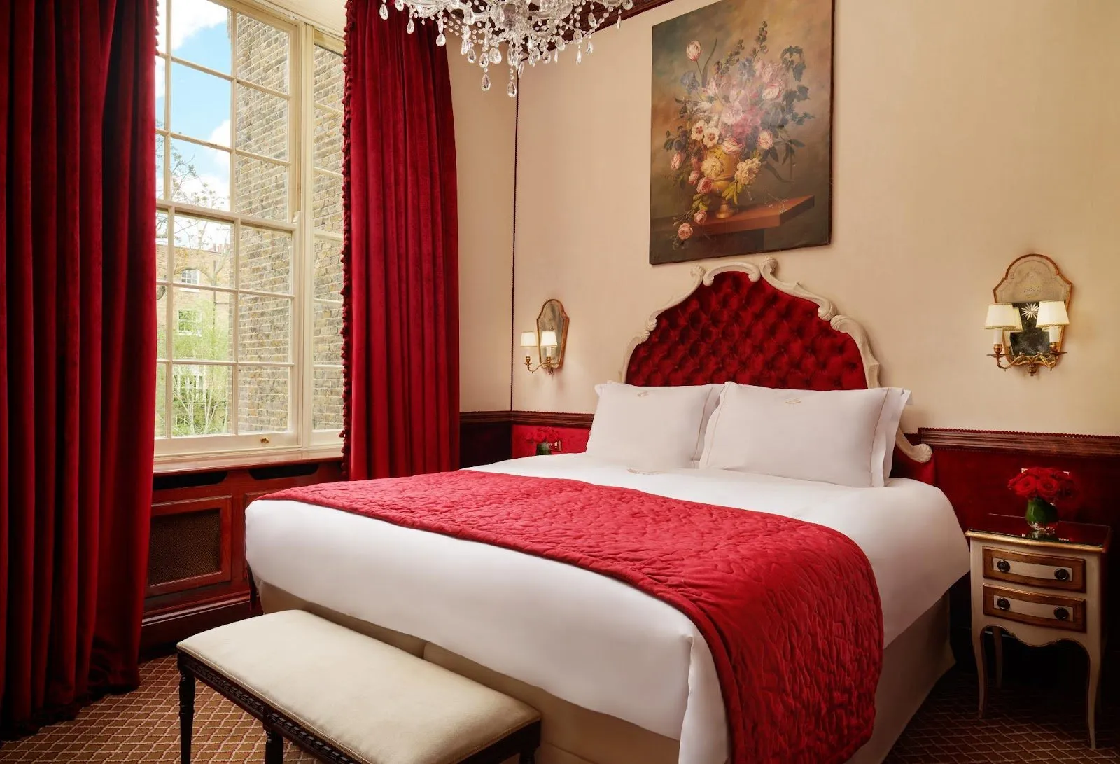 A red-themed bedroom at The Montague on the Gardens, with a crystal chandelier, a still-life painting and a sash window over a brick townhouse