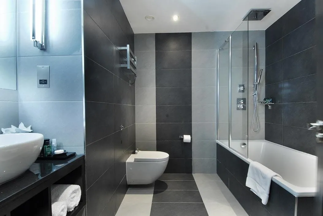 A bathroom at Montcalm Brewery in slate-grey and pale grey tiles, with a shower over the bath behind a glass screen, a wall-hung toilet and a bowl basin on a black counter