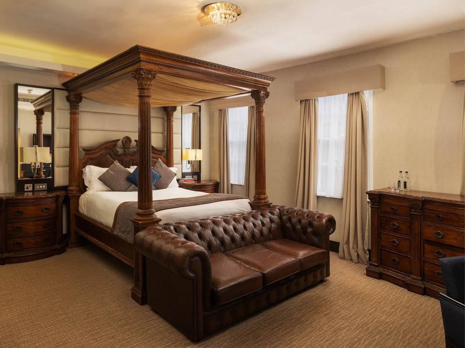 The Montcalm Residence at Montcalm Brewery: a carved four-poster bed with a draped canopy, a brown leather Chesterfield sofa, mahogany chests of drawers and two sash windows with cream curtains