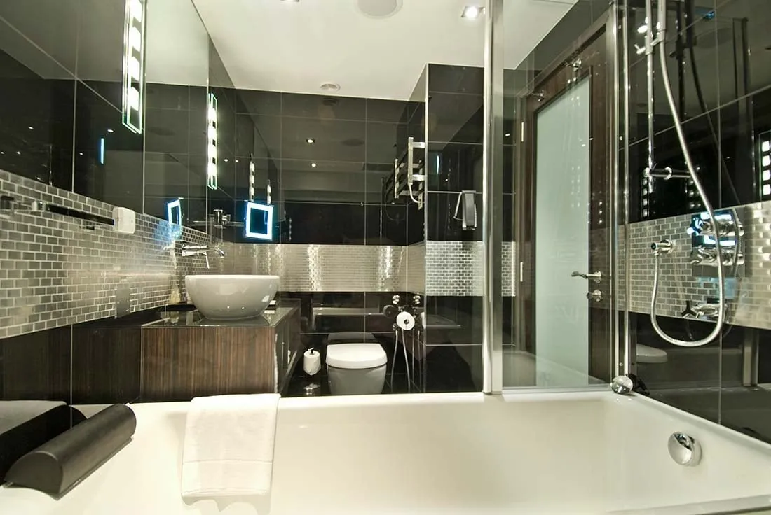 A Montcalm Mayfair bathroom in black glass and silver mosaic tiles, with a white bowl basin on a dark wood counter and a bath with a glass shower screen in the foreground