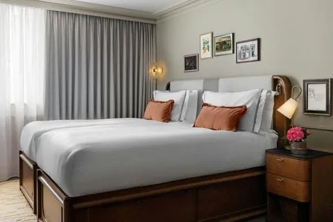 A bedroom at Montcalm Mayfair with a wide bed on a dark wooden base, rust-coloured cushions, framed prints above the headboard and grey curtains over sheers at the window