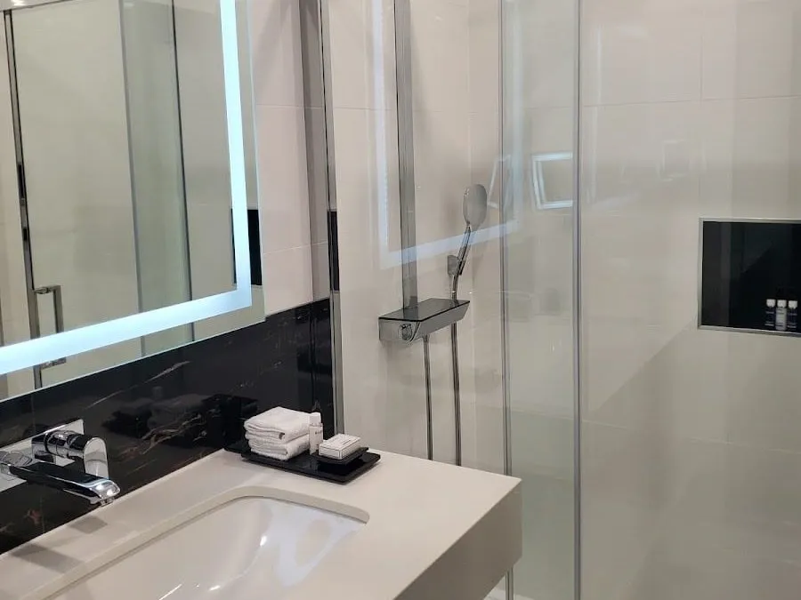 A bathroom at Montcalm Royal London House with a walk-in shower behind a glass screen, cream tiles, a black marble splashback and a backlit mirror over the basin