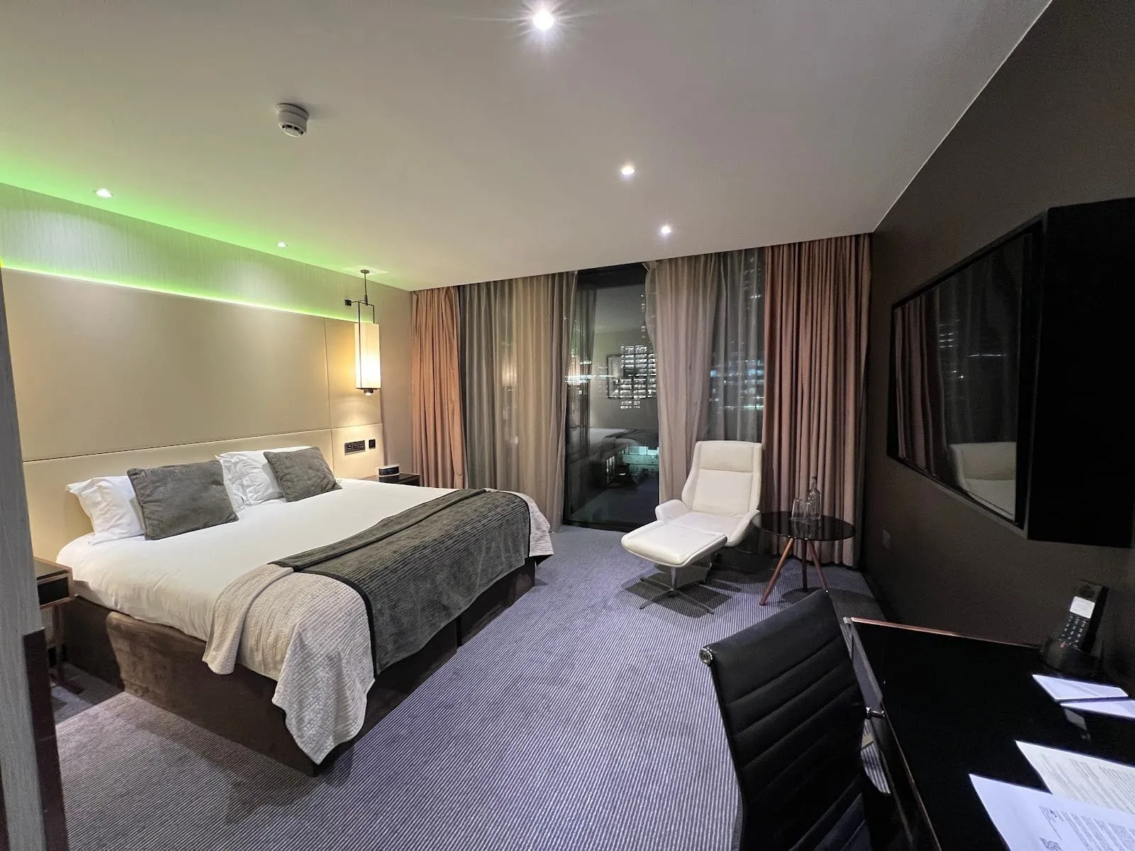A king room at Montcalm Royal London House at night, with a green-lit headboard wall, a grey throw, a white lounge chair and footstool, and a floor-to-ceiling window onto lit office towers