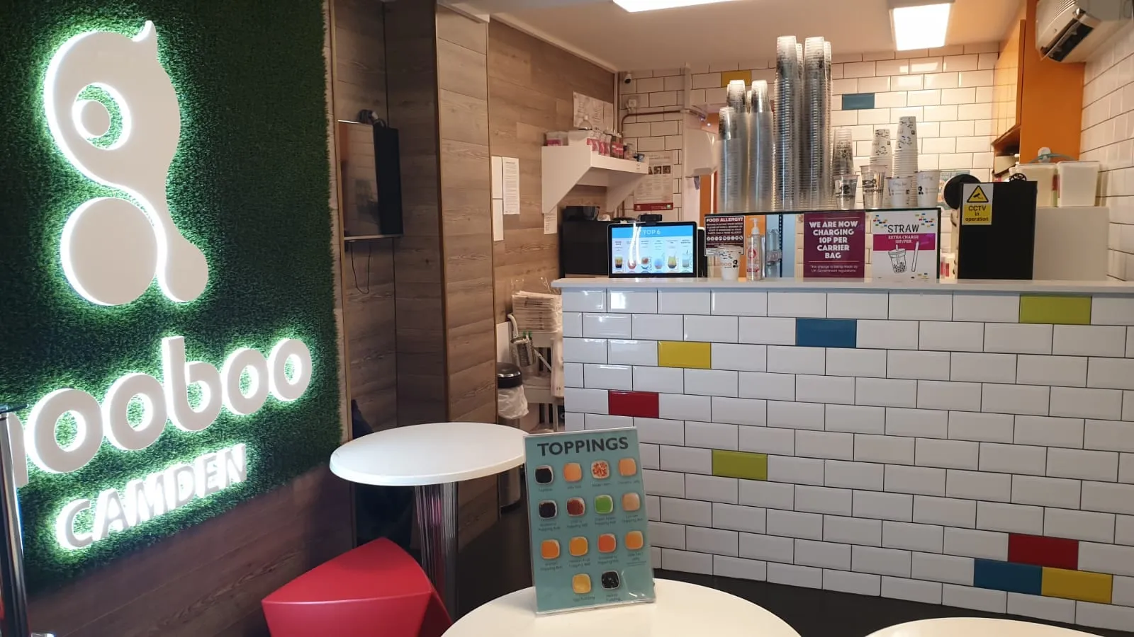 Inside Mooboo Camden: a lit Mooboo logo on a green wall, a white tiled counter and a toppings menu on a small table