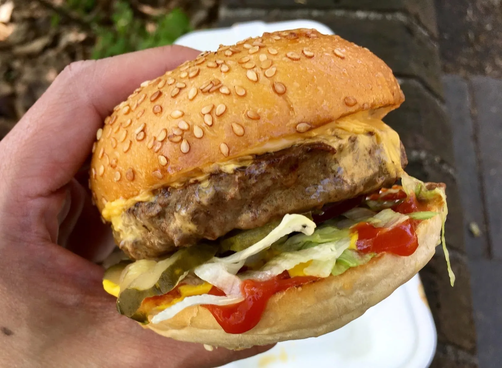 A Mother Flipper cheeseburger held in one hand: a thick beef patty with melted American cheese, pickles, shredded lettuce, ketchup and mustard in a glossy sesame bun