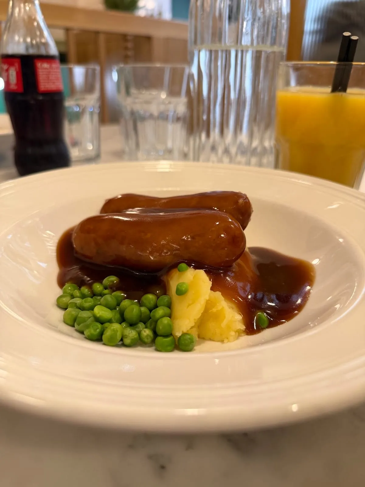 Two sausages under onion gravy with mashed potato and peas, on a white plate.
