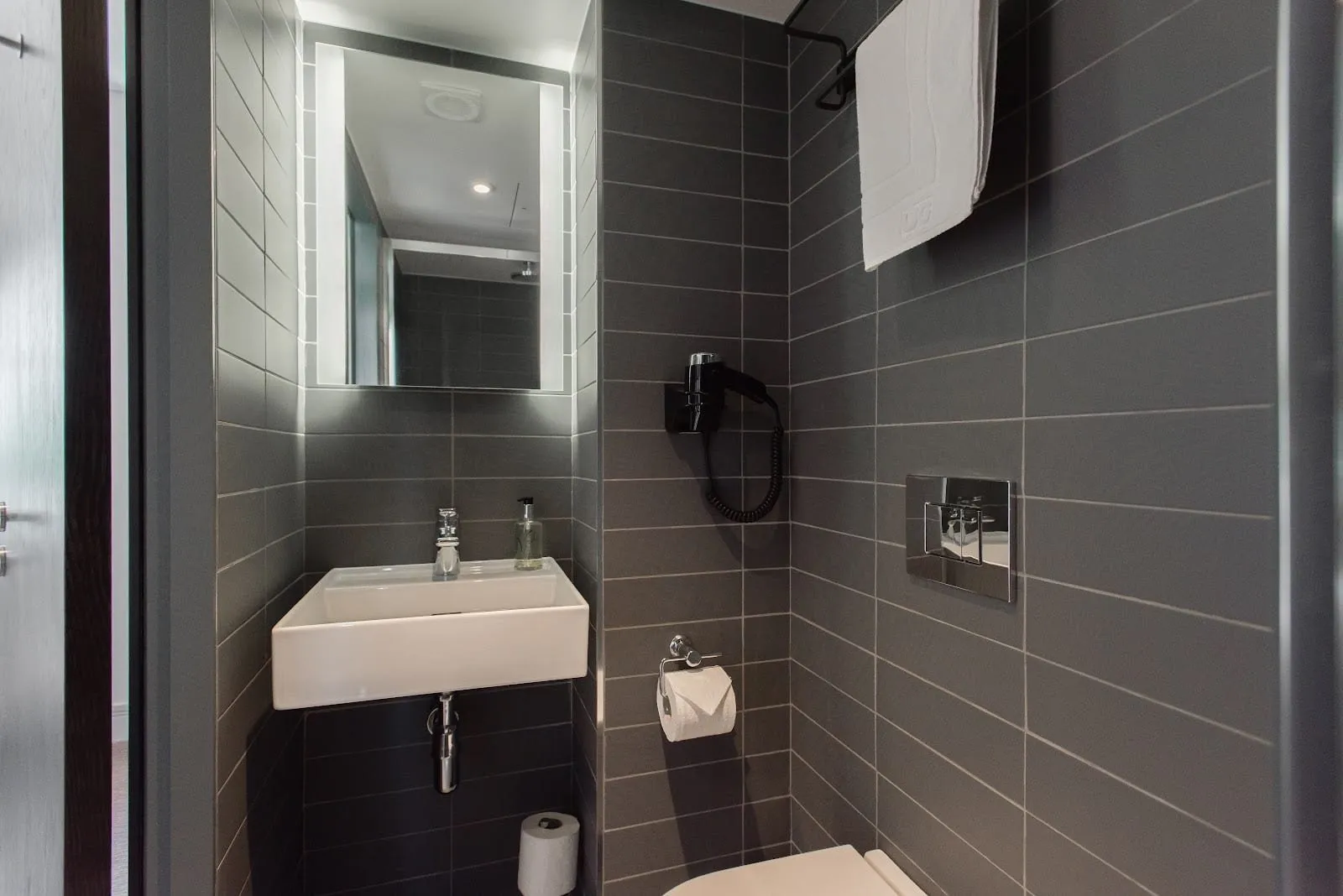 A compact shower room at Mowbray Court in dark grey tiles, with a small wall-hung basin, a lit mirror, a wall-mounted hairdryer and a towel shelf