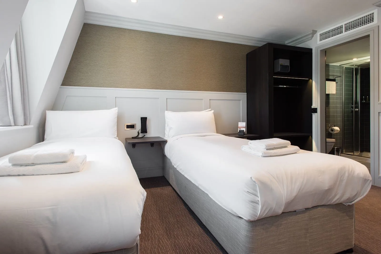An attic twin room at Mowbray Court with two single beds under a sloping ceiling, white panelling, an open dark wardrobe and an air-conditioning grille, with the shower room through the door