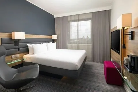 A double room at Moxy London Stratford with a padded charcoal headboard on a wood strip, white-shaded wall lamps, a grey swivel chair, a magenta pouffe and a window looking over trees and distant towers