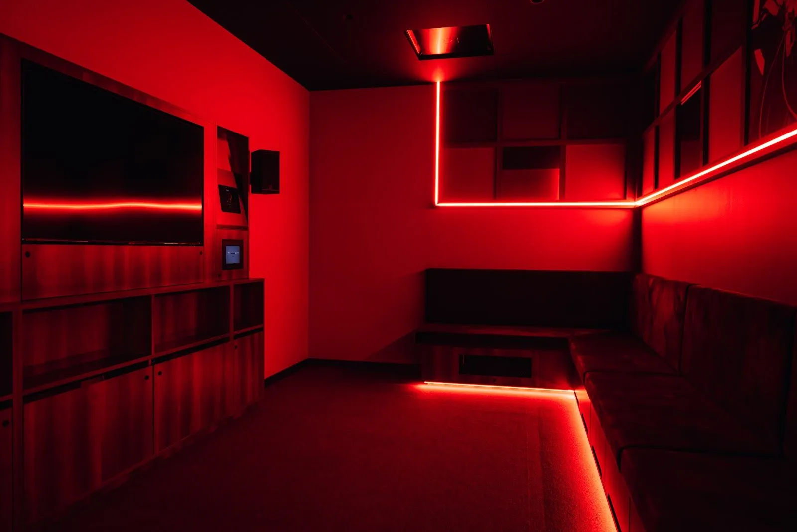 A Moyagi karaoke room lit red, with a wall-mounted screen, a control panel and a banquette along one wall
