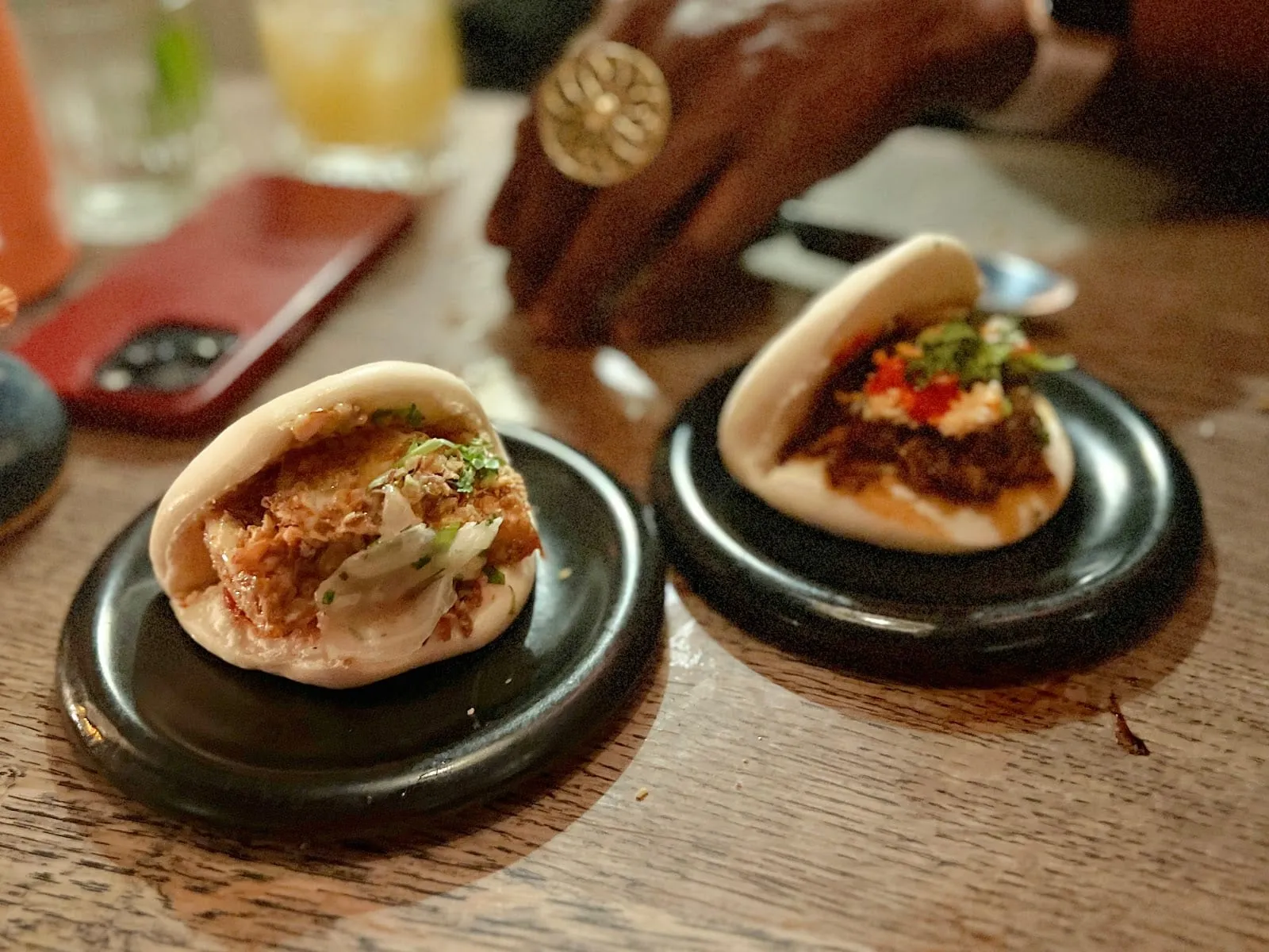 Two steamed bao buns on small plates, one filled with a crispy fried filling and shredded slaw, the other with braised meat, chilli and coriander