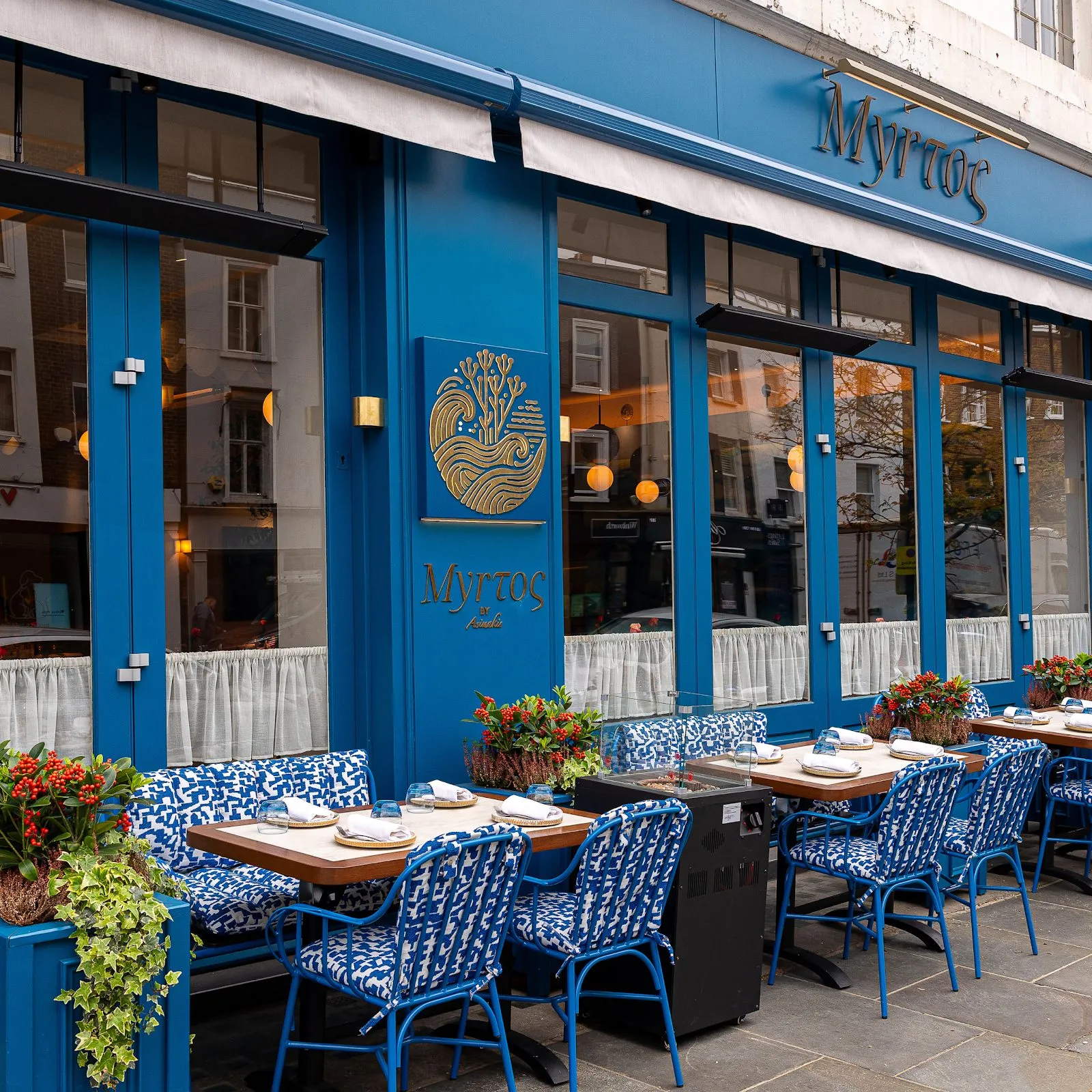 Myrtos's blue shopfront on Brompton Road, its name in Greek letters above the windows, with blue-and-white patterned chairs at pavement tables