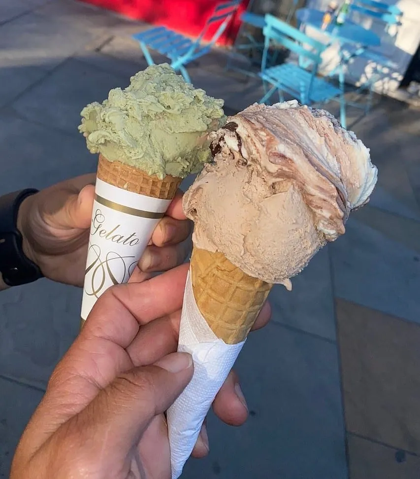 Two cones held over the pavement outside the shop, one pistachio and one a chocolate and cream swirl, with blue café chairs behind