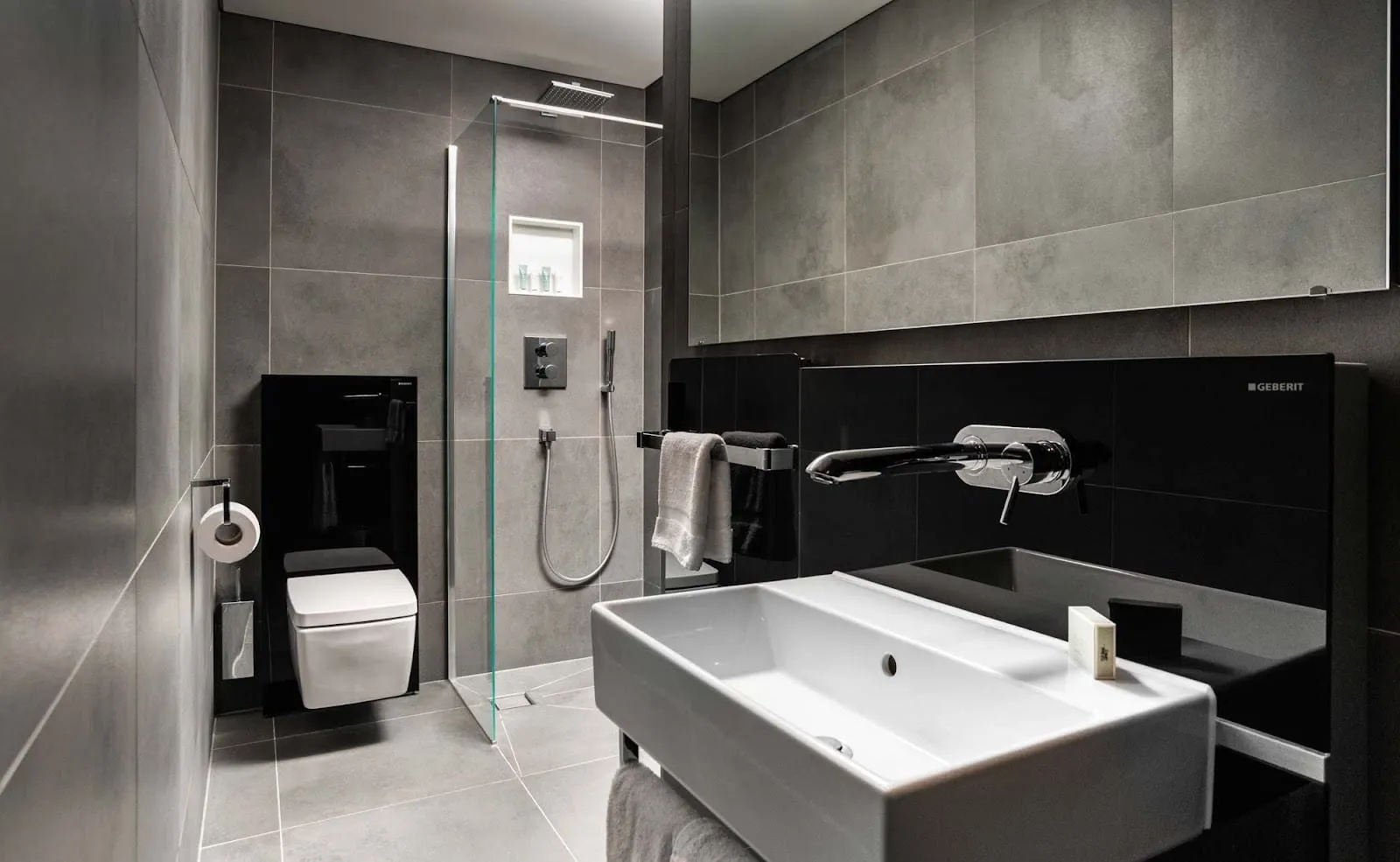 A bathroom at Native Bankside with grey stone-effect tiles, a walk-in rain shower behind a glass panel, a wall-hung toilet and a wide white basin