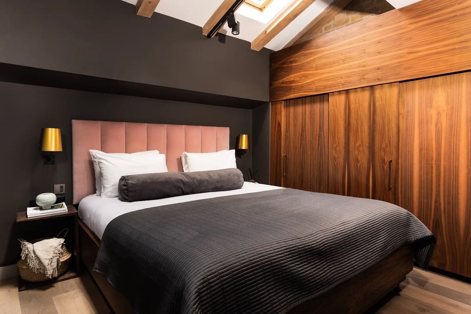 An apartment bedroom at Native Bankside with a pink velvet headboard, charcoal walls, a wall of walnut wardrobes, exposed timber roof beams, a skylight and bare brick