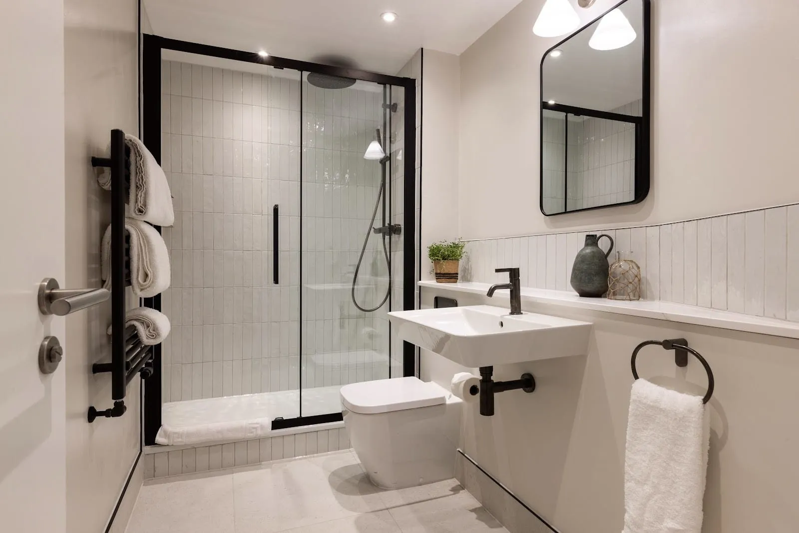 A bathroom at Native Hyde Park with a black-framed glass walk-in shower, white vertical tiles, a wall-hung basin with black taps and a black heated towel rail