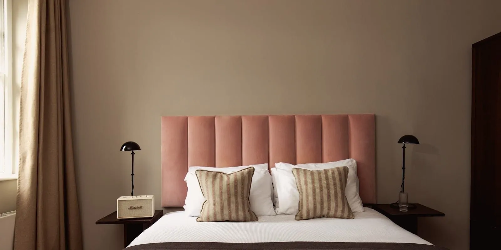 A double bed at Native Hyde Park with a dusty-pink velvet panelled headboard, striped cushions, black mushroom lamps on dark side tables and a tall sash window with linen curtains