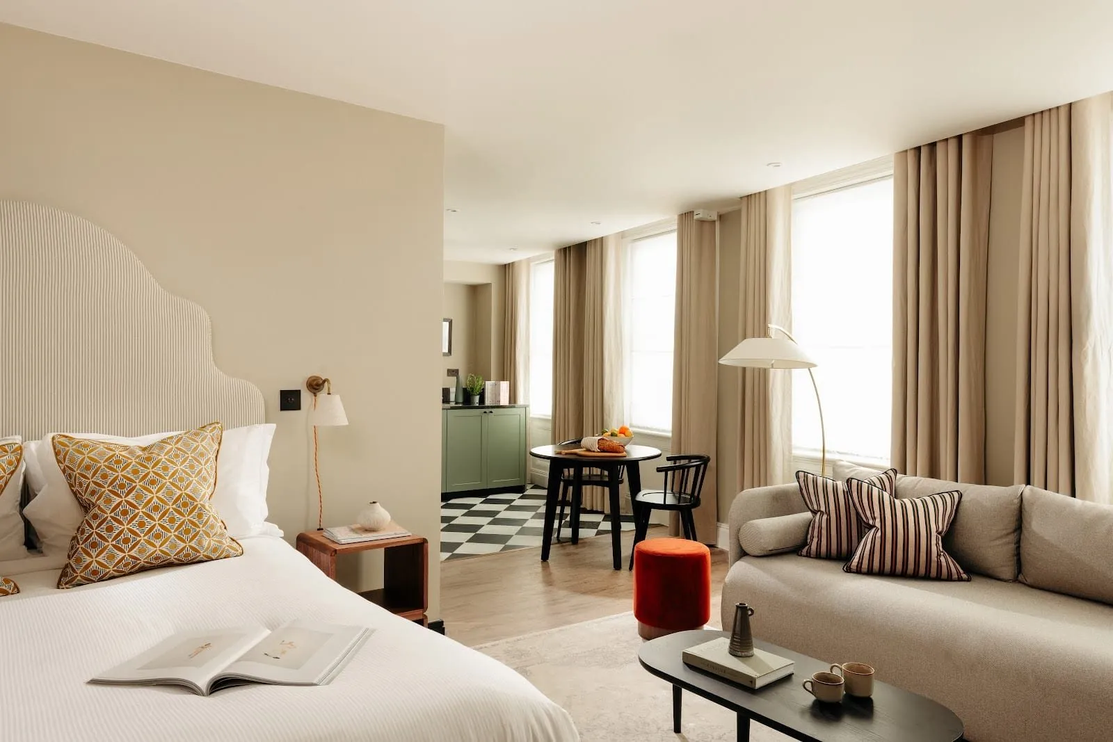 A studio at Native King's Wardrobe with a scalloped striped headboard and mustard patterned cushions, a grey sofa and orange footstool, tall linen-curtained windows, and a sage-green kitchen and round table on a black-and-white chequered floor beyond