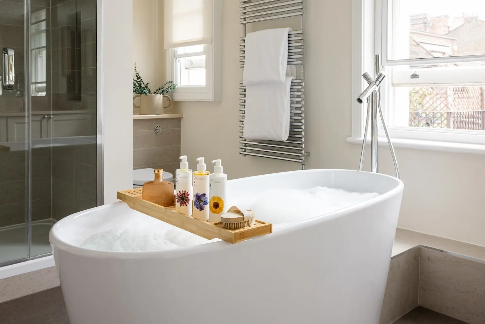 A Native Mayfair bathroom with a freestanding oval bath under a sash window, a floor-standing chrome filler, a heated towel rail and a separate glass-walled shower