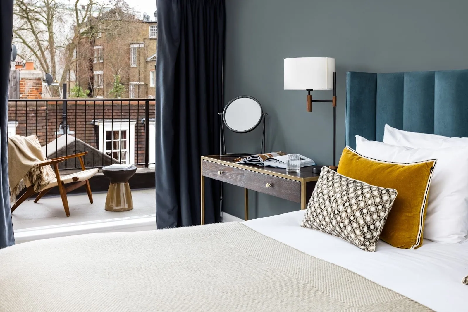 A Native Mayfair bedroom with a teal velvet headboard, mustard and patterned cushions and a dressing table with a round mirror, beside a door open onto a roof terrace with a wooden armchair