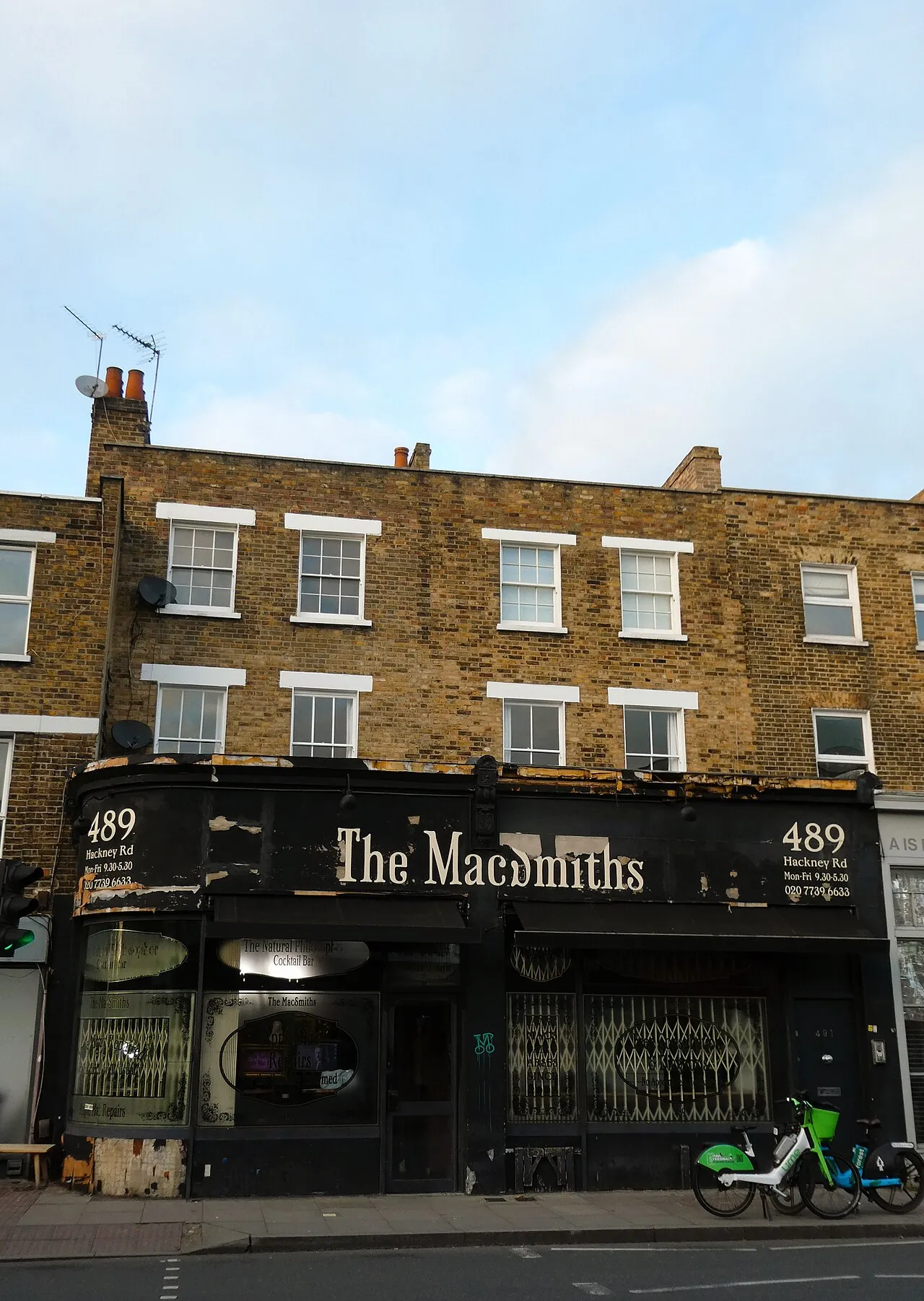 A black shopfront signed The MacSmiths, 489 Hackney Rd, with a small Natural Philosopher cocktail bar sign in the window, under a brick terrace