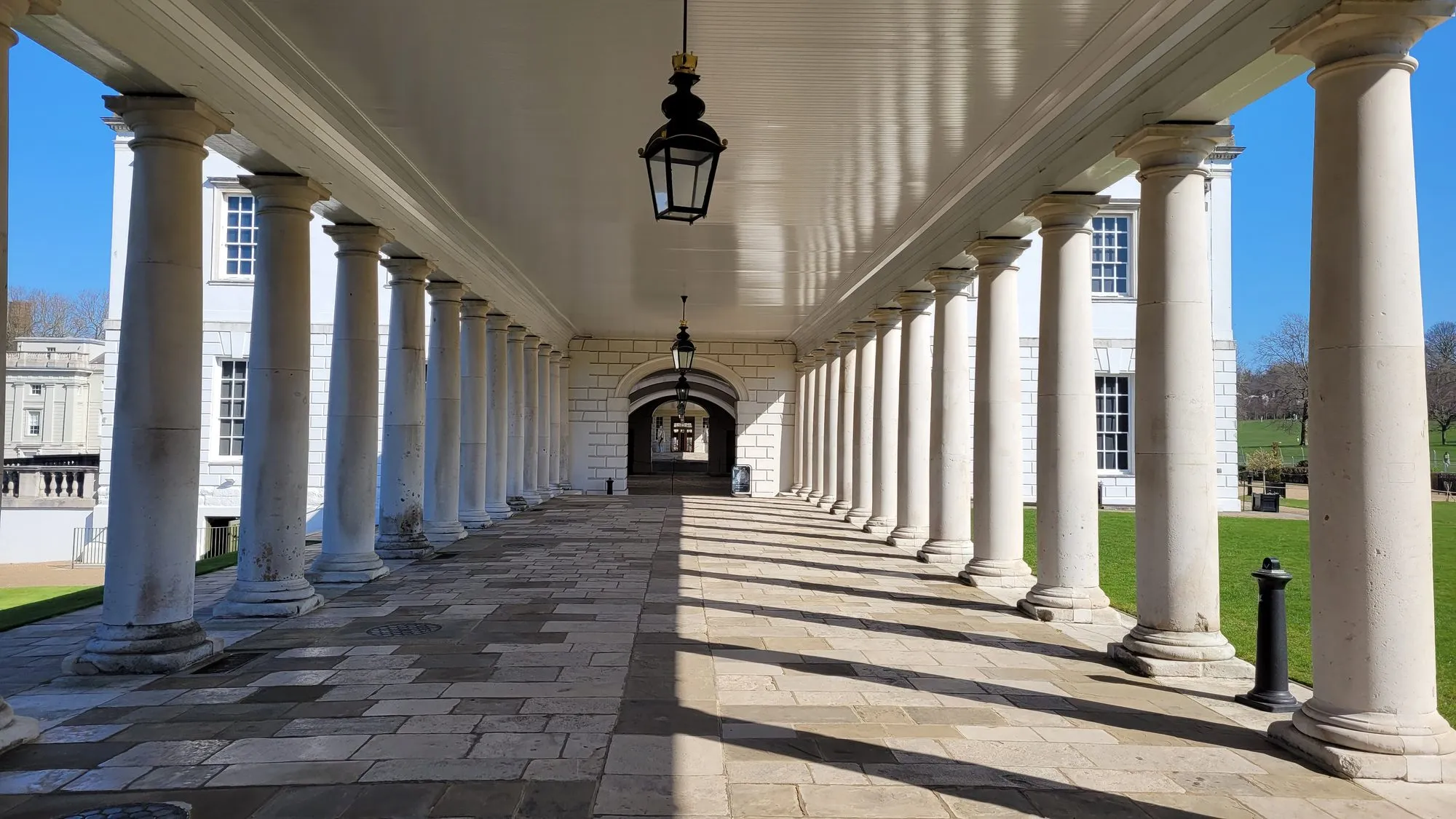 The colonnade beside the Queen's House at the Old Royal Naval College, its white columns throwing long shadows across the flagstones