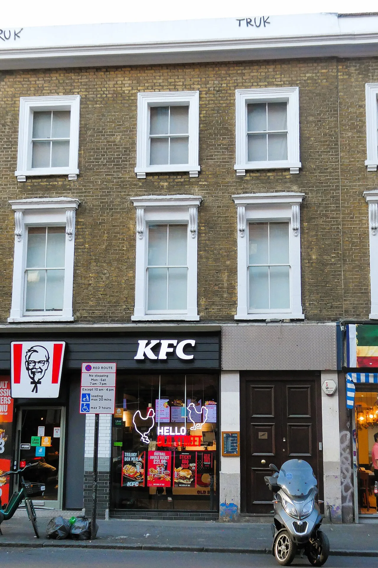 A plain pair of dark wooden doors with a small brass bird plaque, beside a KFC on City Road, under three floors of yellow-brick flats