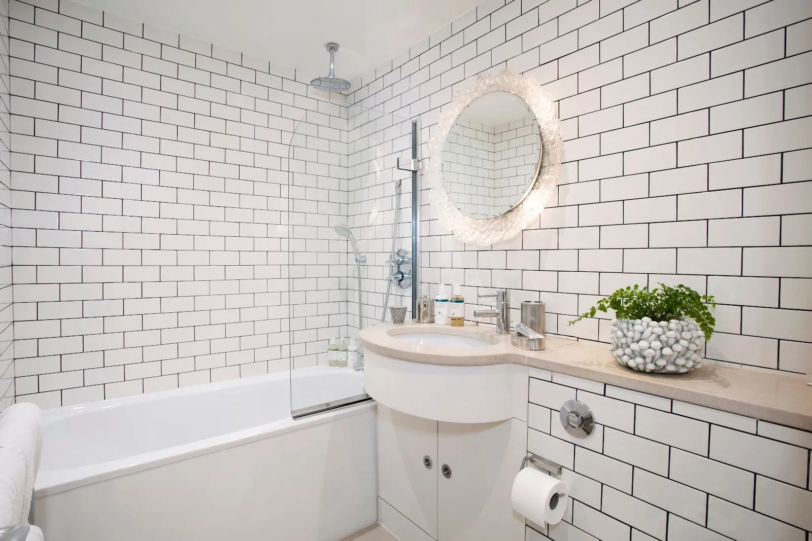 A No.5 Maddox Street bathroom in white metro tiles, with a bath under a glass screen and ceiling rainfall shower, and a curved vanity below a round glass-framed mirror