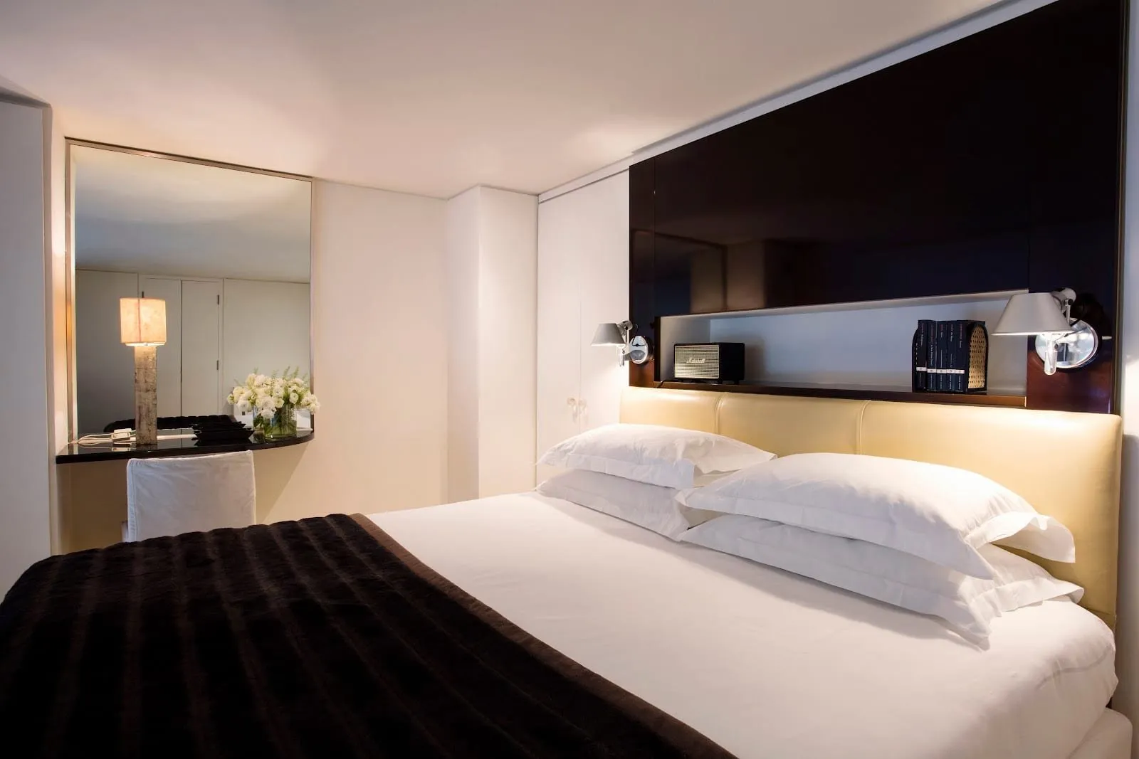 A No.5 Maddox Street bedroom with a cream leather headboard beneath a black lacquered panel, a lit shelf holding a Marshall speaker and books, and a dressing table with a tall mirror