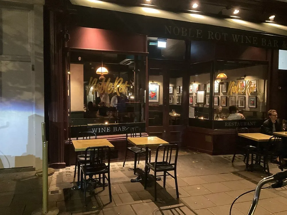 The dark green and gold frontage of Noble Rot at night, lettered Noble Rot Wine Bar, with WINE BAR etched on one window and RESTAURANT on the other and tables on the pavement