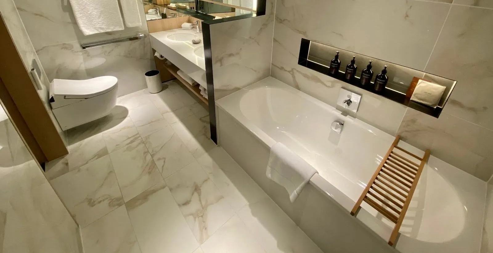 A Nobu Portman Square bathroom in white marble-effect tiles with a deep bath, a lit niche of toiletries, a double basin behind a partition and a wall-hung Japanese-style toilet