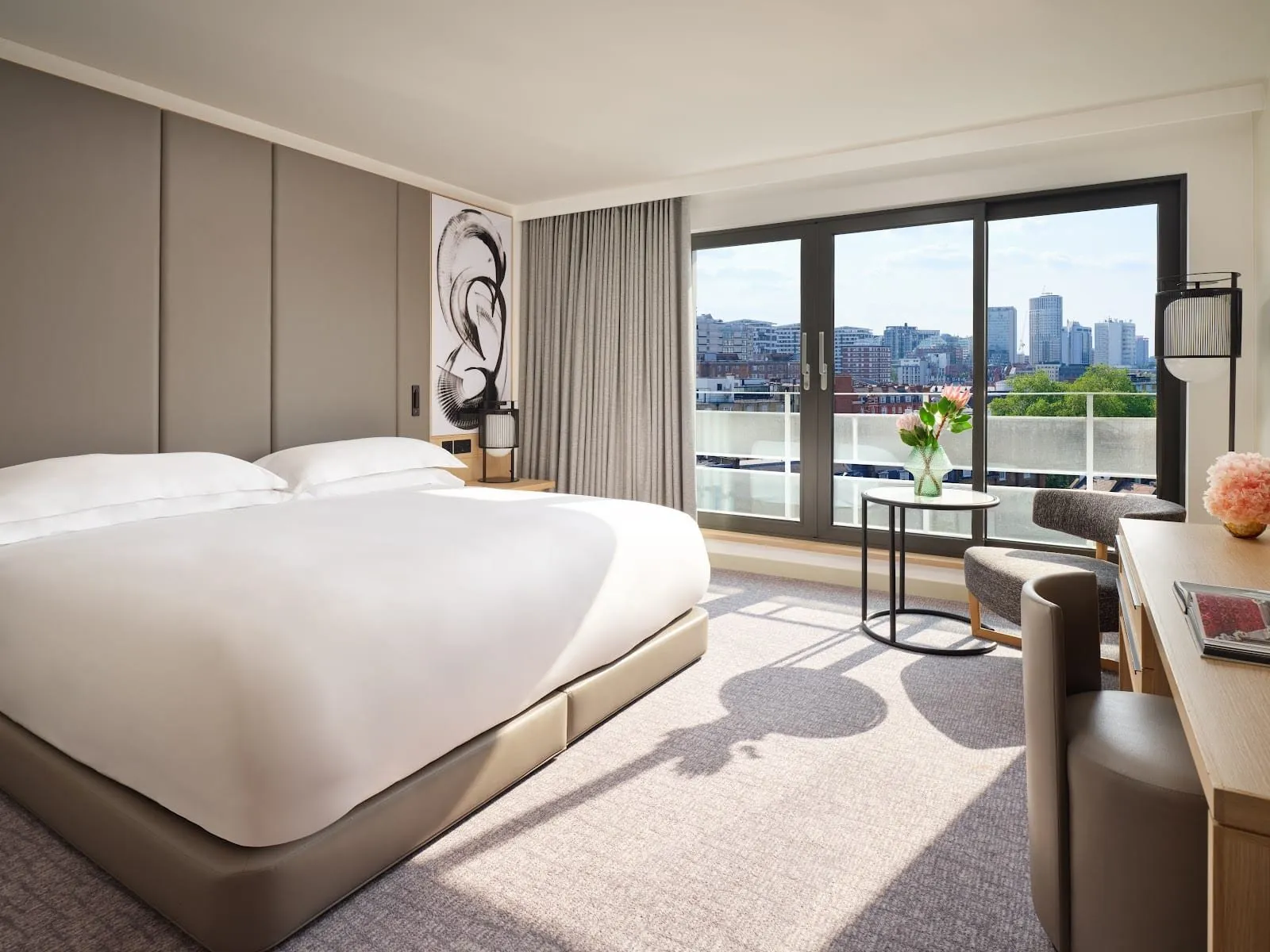 A Nobu Portman Square bedroom with a wide bed against a padded taupe wall, a black ink-brush artwork, and floor-to-ceiling glass doors opening on to a balcony with a view over rooftops to tower blocks