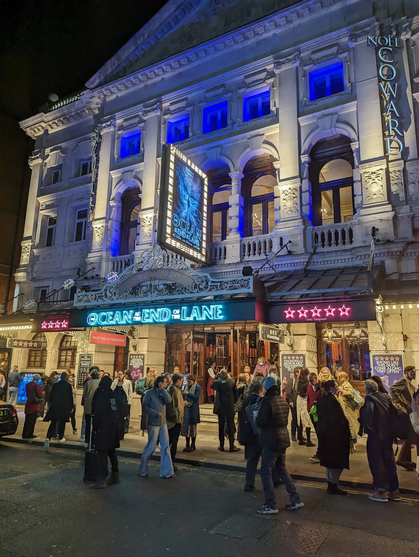 The lit facade of the Noel Coward Theatre at night, with an audience gathering outside before a performance