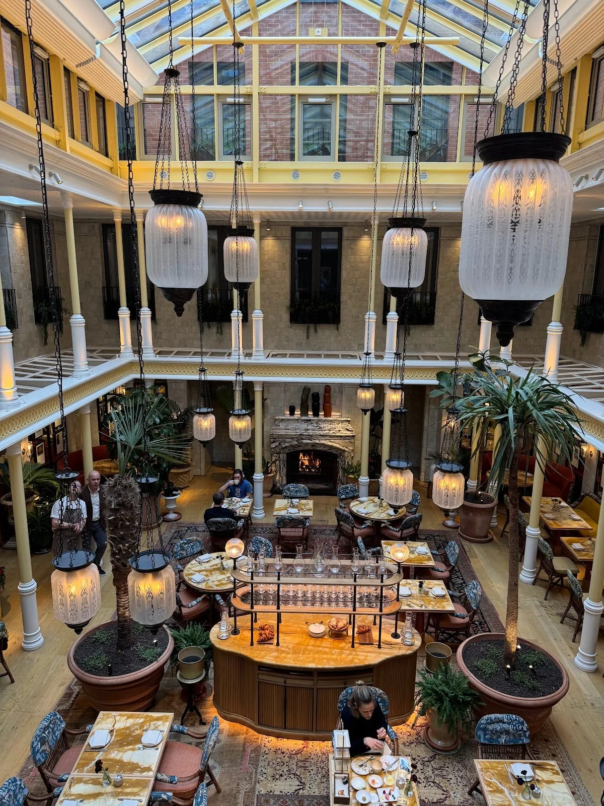 The glass-roofed atrium at NoMad London seen from an upper gallery, hanging lanterns and palms over the tables and a central bar