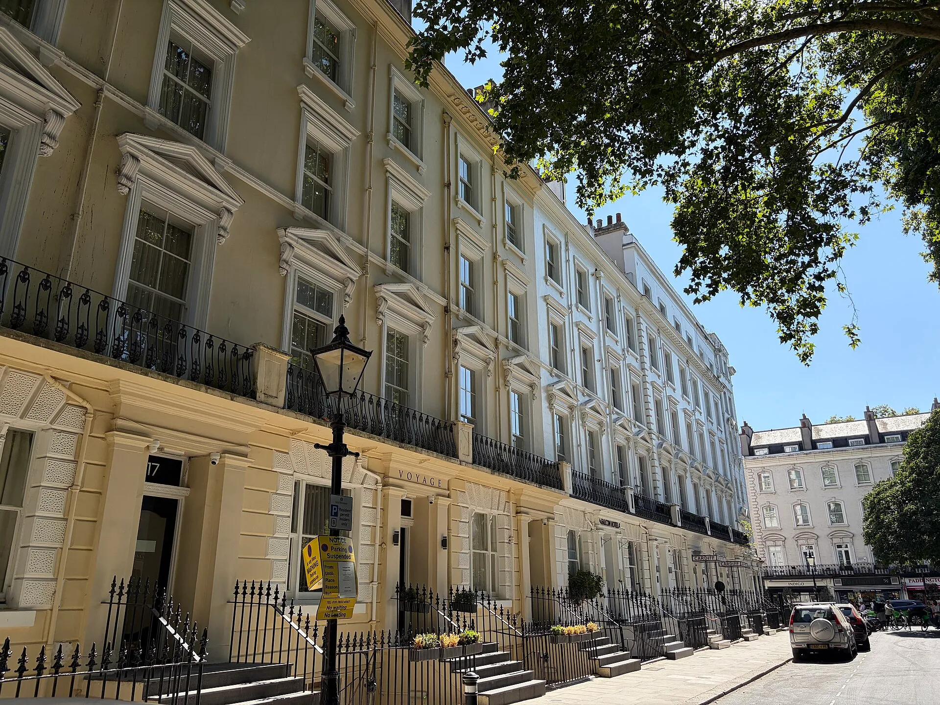 A terrace of cream and white stucco townhouse hotels on Norfolk Square in Paddington, with steps up to each front door behind black railings, and the Hotel Indigo sign on London Street at the far end