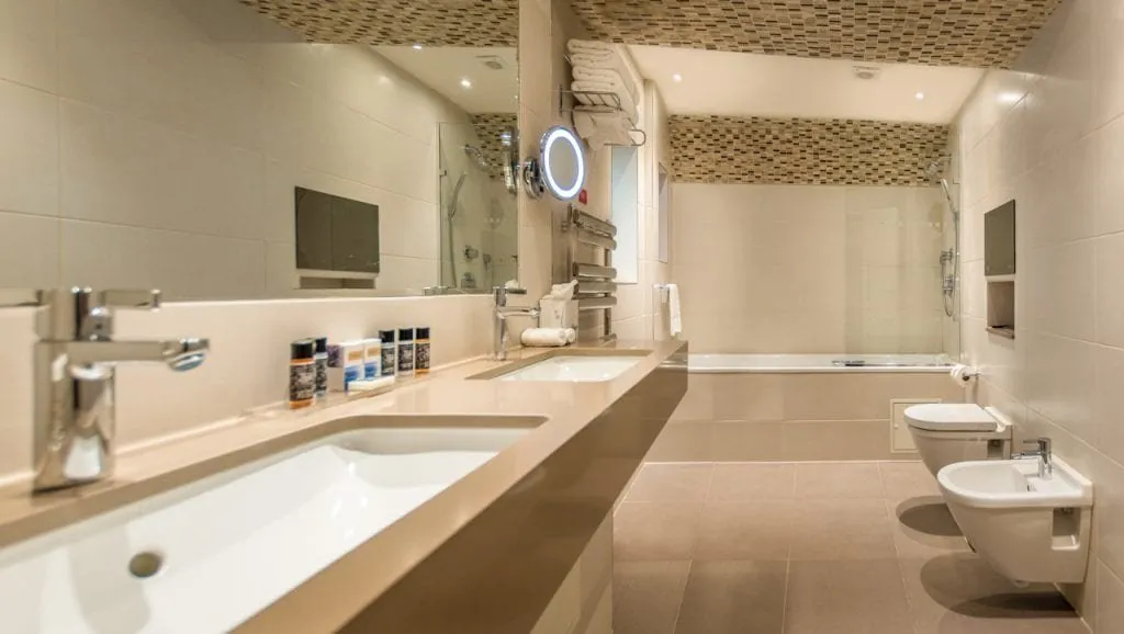 A long beige bathroom at Norfolk Towers Paddington with two basins in a stone counter, a bath with a hand shower at the far end, a wall-hung toilet and a bidet