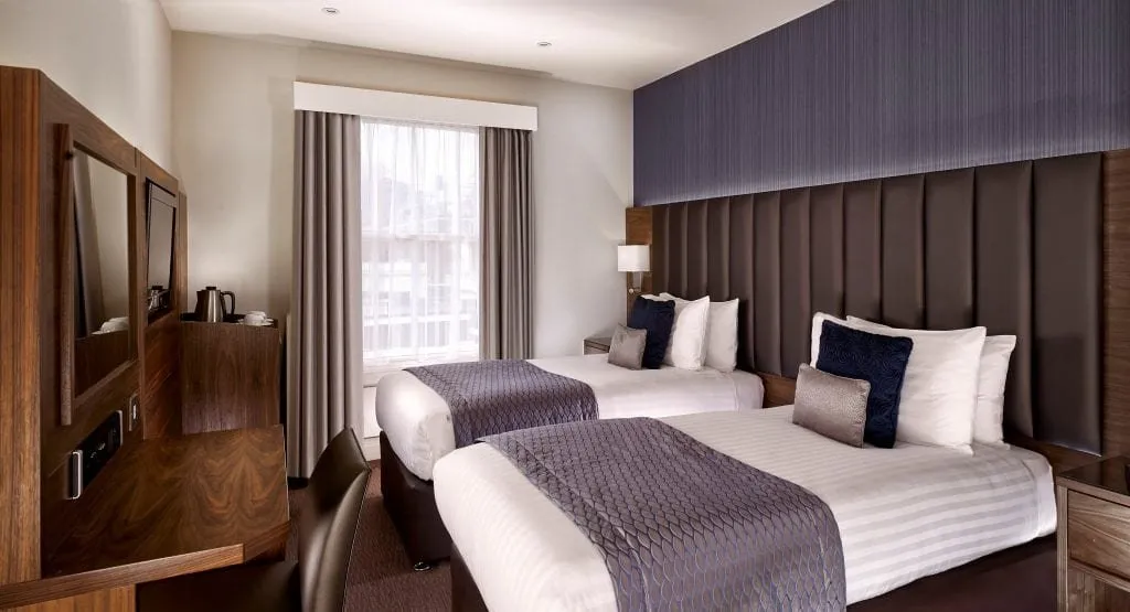 A room at Norfolk Towers Paddington with two double beds under grey-lilac runners, a tall padded headboard wall, a sash window with voile curtains and a walnut desk