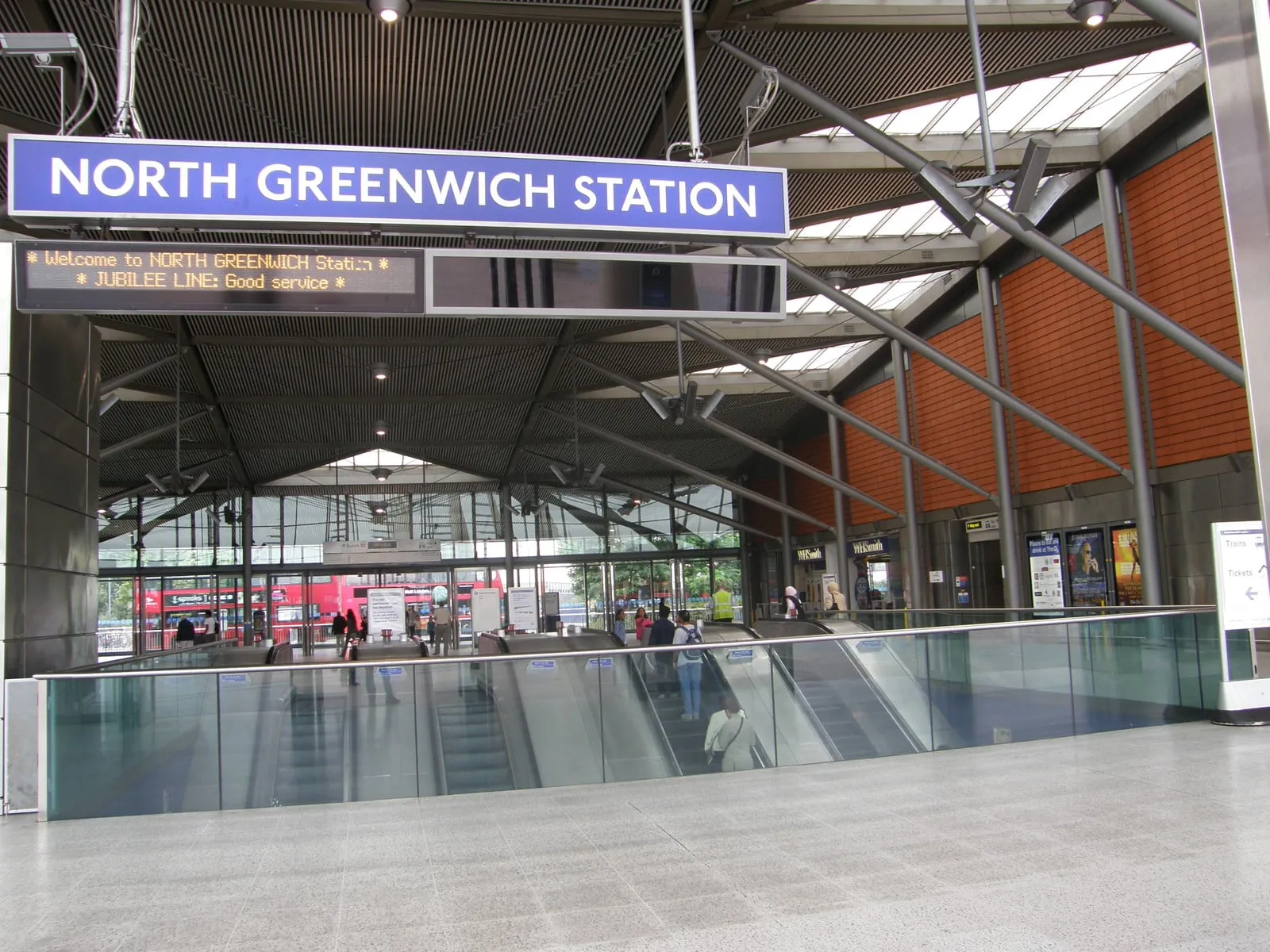 The North Greenwich Station sign above the concourse, with escalators running down behind a glass balustrade and buses visible through the glass doors beyond