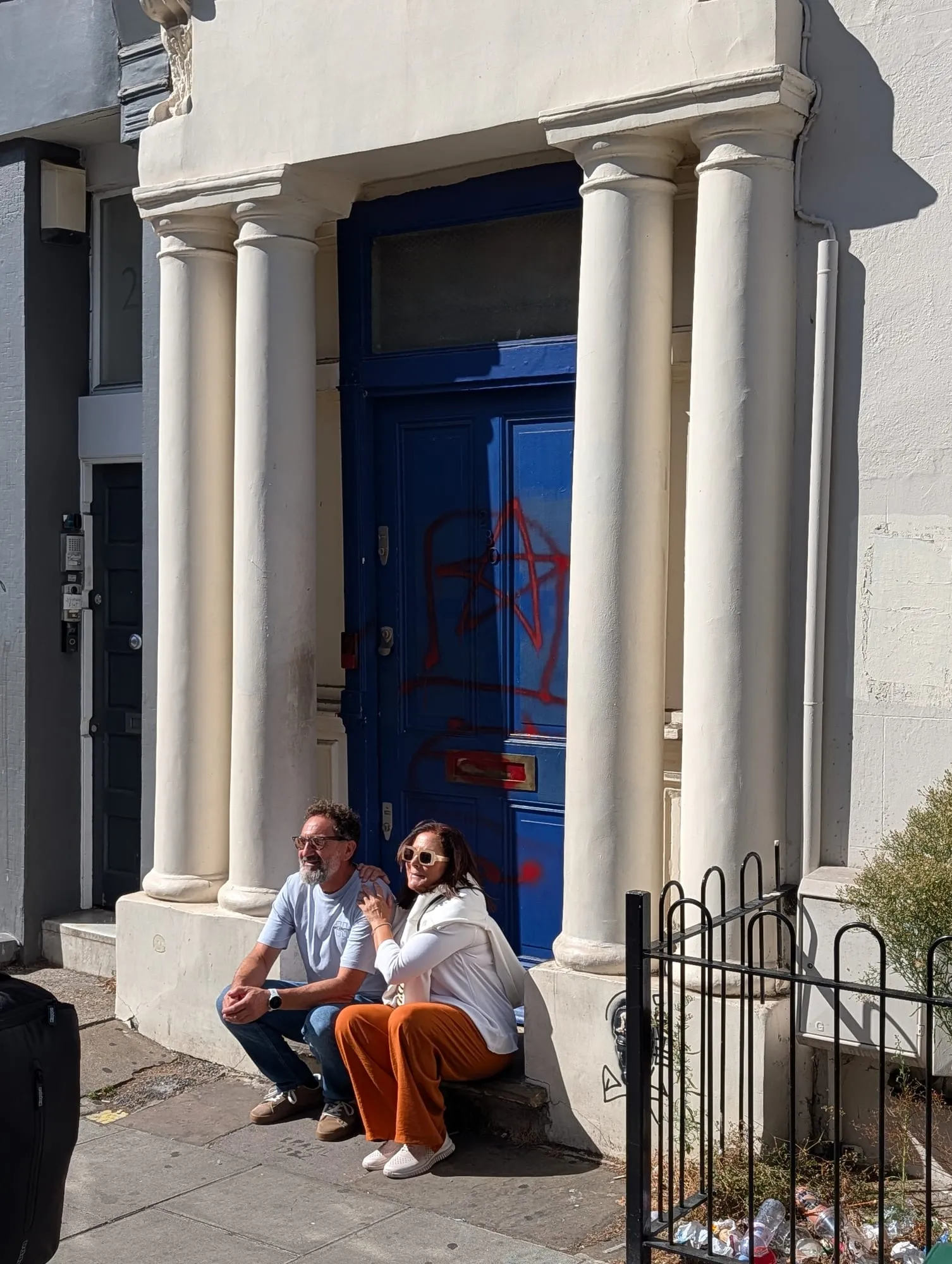 The blue door at 280 Westbourne Park Road between two white columns, defaced with red spray paint, with two visitors sitting on the step