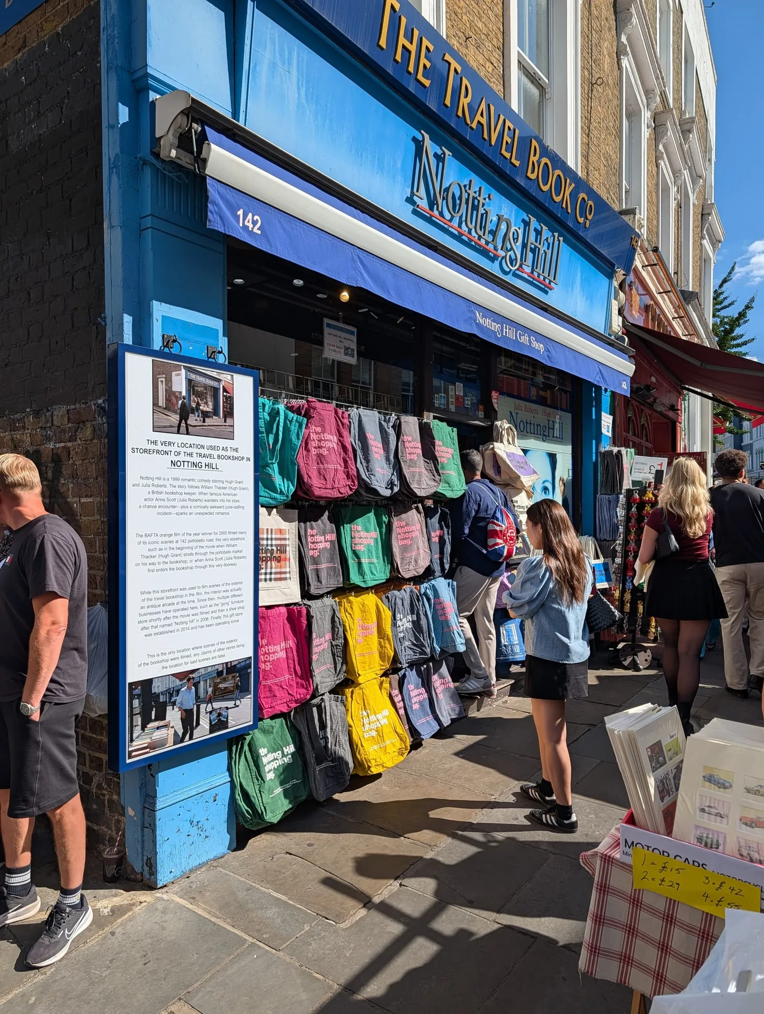 The blue shopfront at 142 Portobello Road painted THE TRAVEL BOOK Co, hung with souvenir tote bags, beside a history board about the film