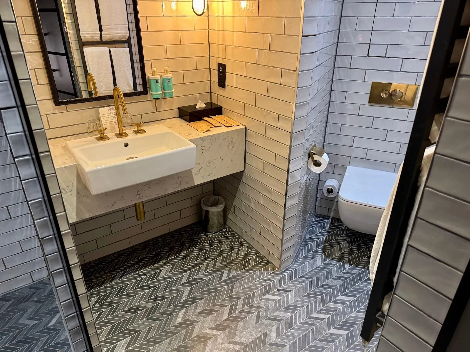 The bathroom at Novotel London Canary Wharf: cream and grey metro tiles, a grey herringbone floor, a basin on a marble-effect shelf with brass taps, and a wall-hung toilet