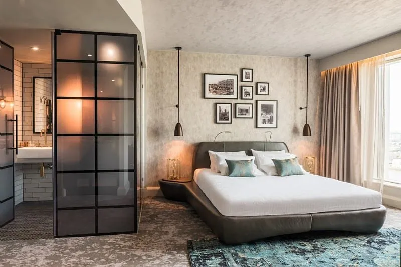 A room at Novotel London Canary Wharf with a low grey leather bed, teal cushions and rug, black-and-white framed prints over the bed, and a black-framed smoked-glass wall around the bathroom