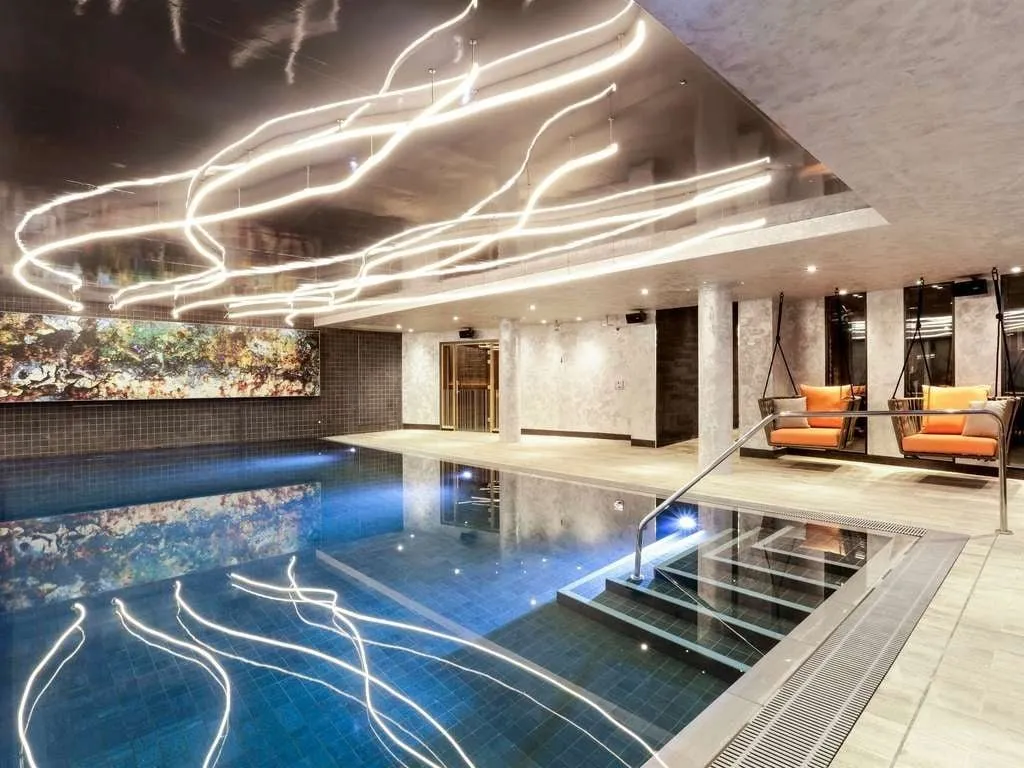 The indoor pool at Novotel London Canary Wharf, with a mural along the far wall and hanging swing chairs